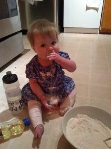 Small child in blue dress and white pants sits next to a bowl of dry ingredients. Their fingers are in their mout as they try edible play dough