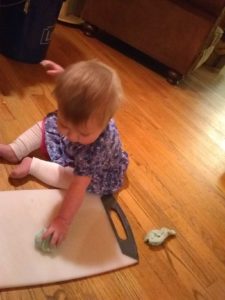 Small child smooshes edible playdough on a cutting board while sitting on the floor.