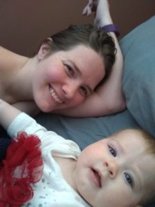 A securely attached mom and baby lay in bed and smile at the camera together