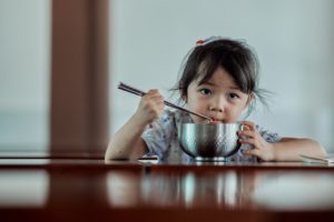 Child sits at table eating a meal from a bowl with chopsticks