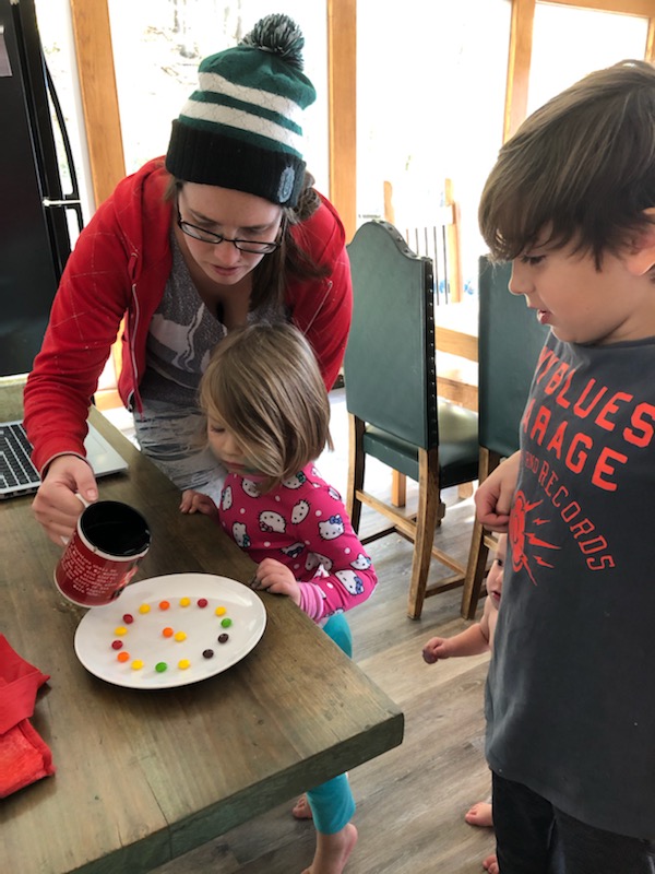 An adult pours water into a plae of skittles while two children watch